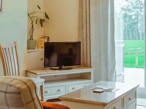 Sala de estar con televisor sobre un mueble blanco, una planta y vista a un paisaje verde a través de una ventana.
