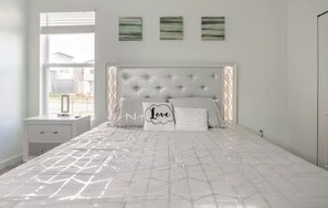 Un dormitorio con un cabecero capitonado, una cama con colcha de patrón geométrico, una mesita de noche con lámpara y tres obras de arte colgadas en la pared.