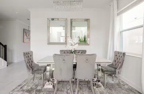 Un comedor con una mesa blanca, cuatro sillas tapizadas, dos espejos en la pared y una araña.