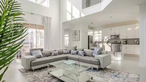 A spacious living room with a large sectional sofa, a glass coffee table, and a modern kitchen in the background. - Spacious 5Bdr 5Bth ChampionsGate Villa Near Disney, Private Pool & Resort Access (Davenport)