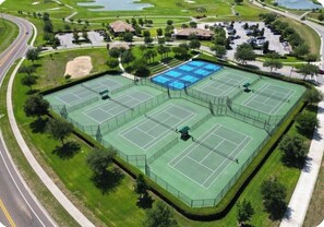 Vue aérienne d’un complexe de courts de tennis comprenant plusieurs terrains, entouré de verdure et doté d’un espace de stationnement.