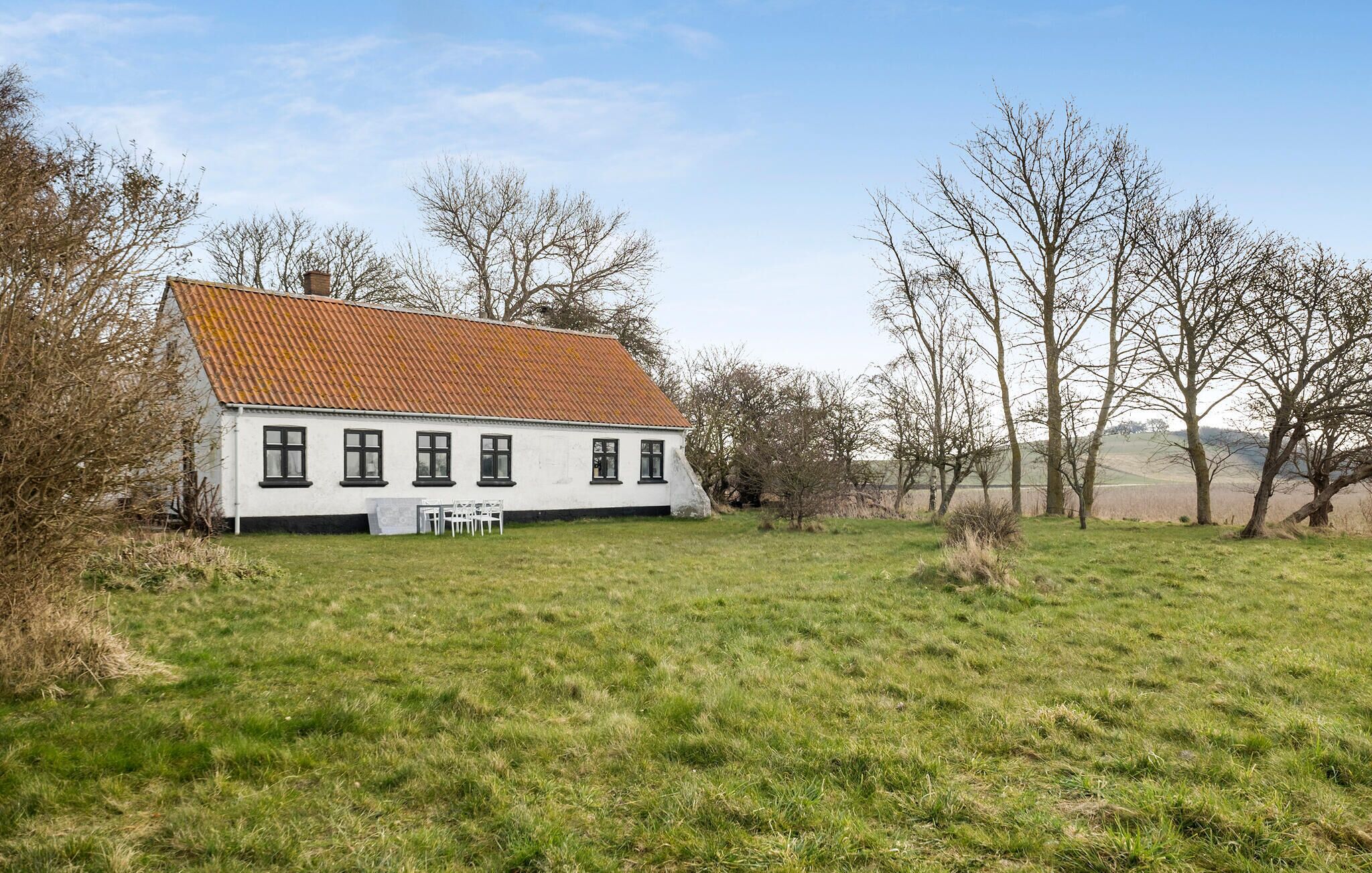 Uma casa branca com telhado de telhas vermelhas, rodeada por um campo gramado e árvores sem folhas.