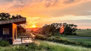 A modern house with a wooden deck and a view of the sunset over a rural landscape.