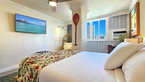 Habitación de hotel con una cama grande, un televisor montado, una silla de ratán, vista de la ciudad a través de la ventana y un ventilador de techo.