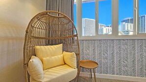 Una silla de mimbre con cojines, una mesita redonda y una ventana con vista a rascacielos.