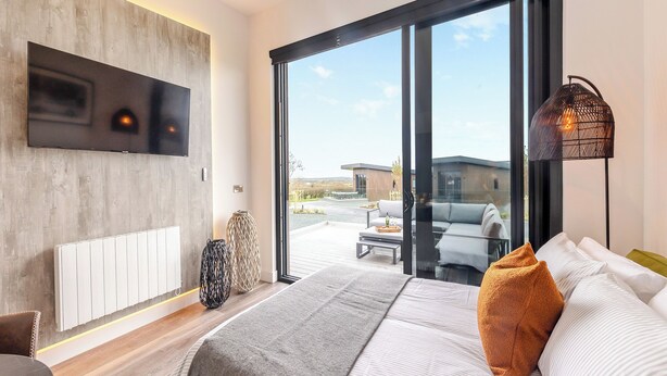 A modern bedroom with a large bed, a TV, a sliding glass door to a balcony, and a floor lamp.