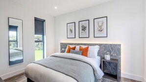 A modern bedroom with a large bed, a nightstand, a mirror, and framed botanical prints on the wall.