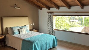 Une chambre avec un plafond en bois, un grand lit recouvert d’une courtepointe turquoise, une table de chevet, une lampe de chevet et une fenêtre avec des rideaux.