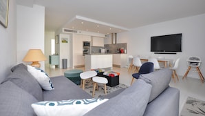 Un salon moderne avec un canapé gris, un coin repas avec une table et des chaises, une cuisine avec des meubles blancs et un téléviseur fixé au mur.