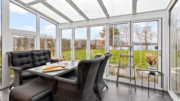 Ein Wintergarten mit Korbmöbeln, einem Tisch mit Brot und Blick auf einen See und Bäume.