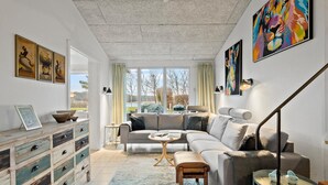 Ein geräumiges Wohnzimmer mit großem Fenster, einem grauen Ecksofa, einer Holzkommode und einem farbenfrohen abstrakten Gemälde.