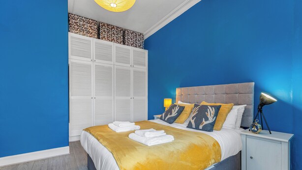 A bedroom with a bed, white wardrobe, and a nightstand with a lamp. - Top Floor Spacious King Size Apartment | City Views | Excellent Transport Links (Edinburgh)