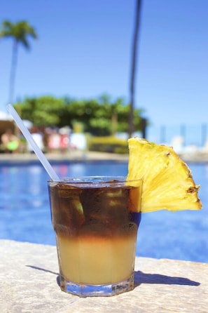 A glass with a layered drink, a straw, and a pineapple slice, placed on a stone surface by a pool with a clear sky.