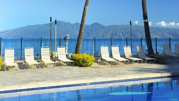 A poolside area with lounge chairs and palm trees, overlooking a clear blue sky and a mountainous backdrop.