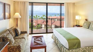 A hotel room with a bed, a sofa, a table, and a balcony with a view of the ocean and buildings.