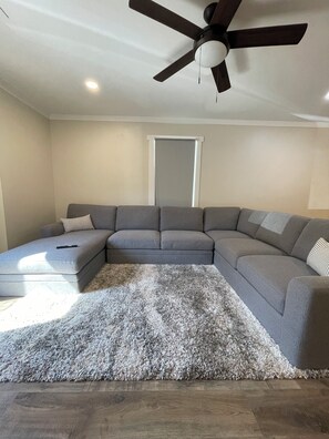 Ein Wohnzimmer mit Ecksofa, einem flauschigen Teppich, Deckenventilator und einem Fenster mit Jalousien.
