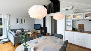 Ein modernes Wohnzimmer mit grauem Sofa, Fernseher und Kamin. Es gibt eine Essecke mit Tisch und Stühlen sowie eine Küche mit weißen Schränken und einer Holz Arbeitsplatte.