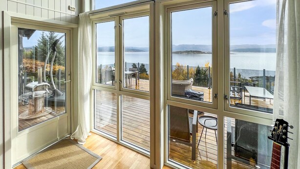 Ein Zimmer mit großen Fenstern mit Seeblick, einer Holzterrasse und einer Gitarre.