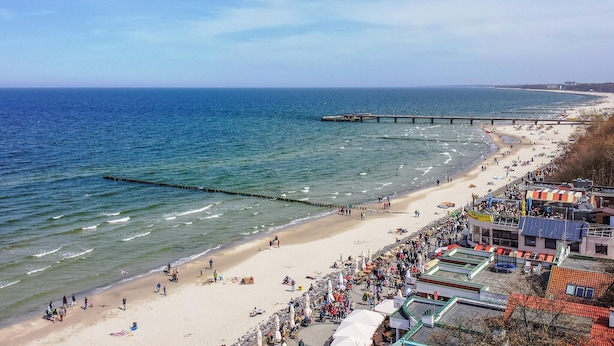 Una playa con un muelle que se adentra en el mar, una orilla arenosa y una multitud de gente.