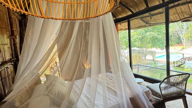 A room with a bed under a canopy, a wicker hanging light fixture, and a view of an outdoor area with a pool. - Akasha suites (Puerto Triunfo)