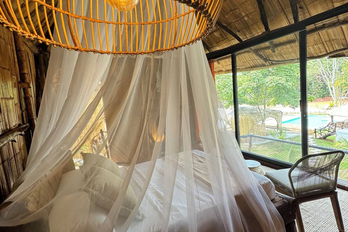 A room with a bed under a canopy, a wicker hanging light fixture, and a view of an outdoor area with a pool.