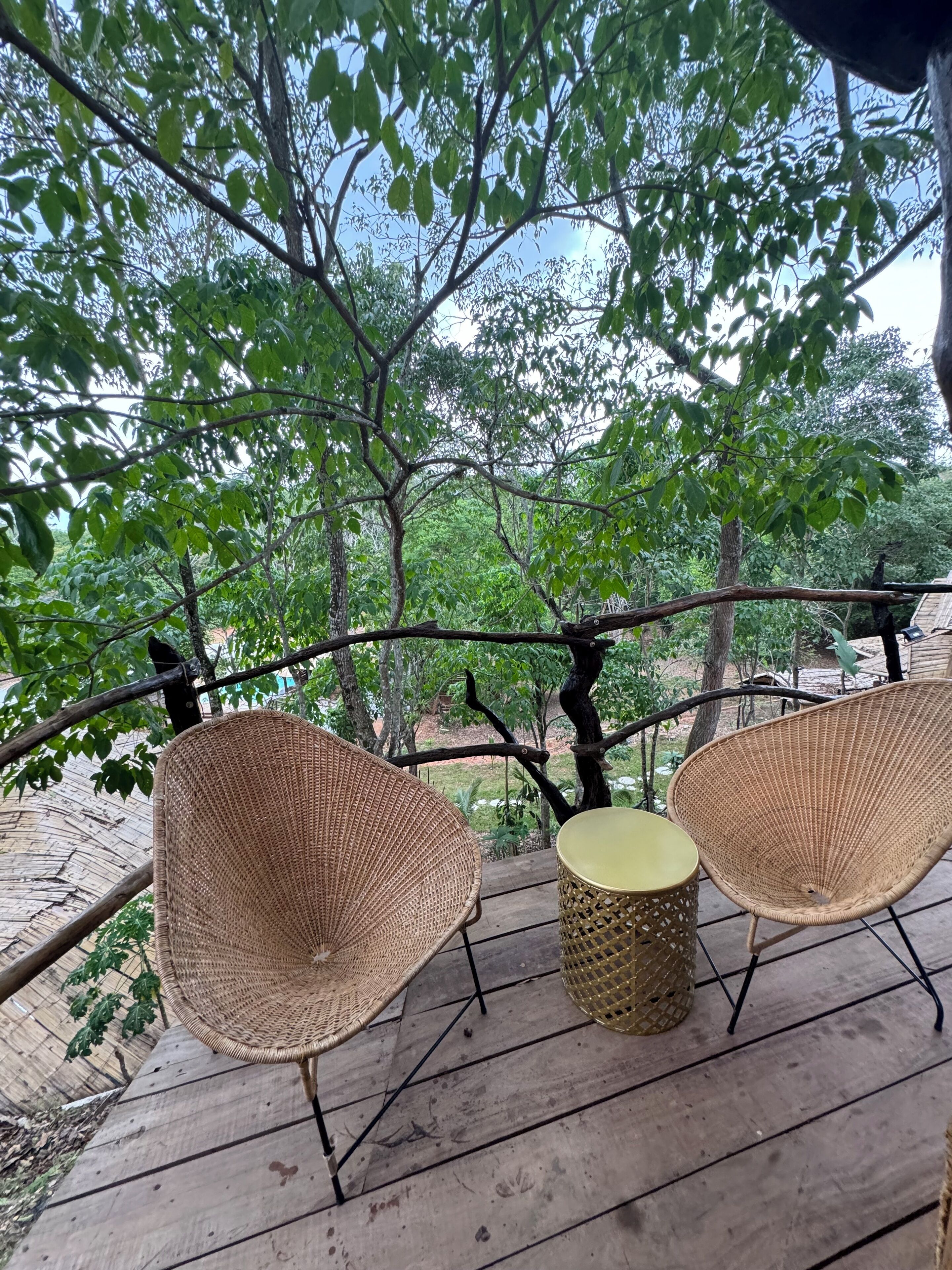 A wooden deck with two woven chairs and a small table, surrounded by trees and a metal railing.