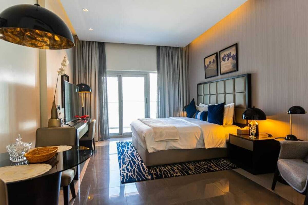 A modern hotel room with a large bed, a desk with a lamp, a chair, and a dining table.