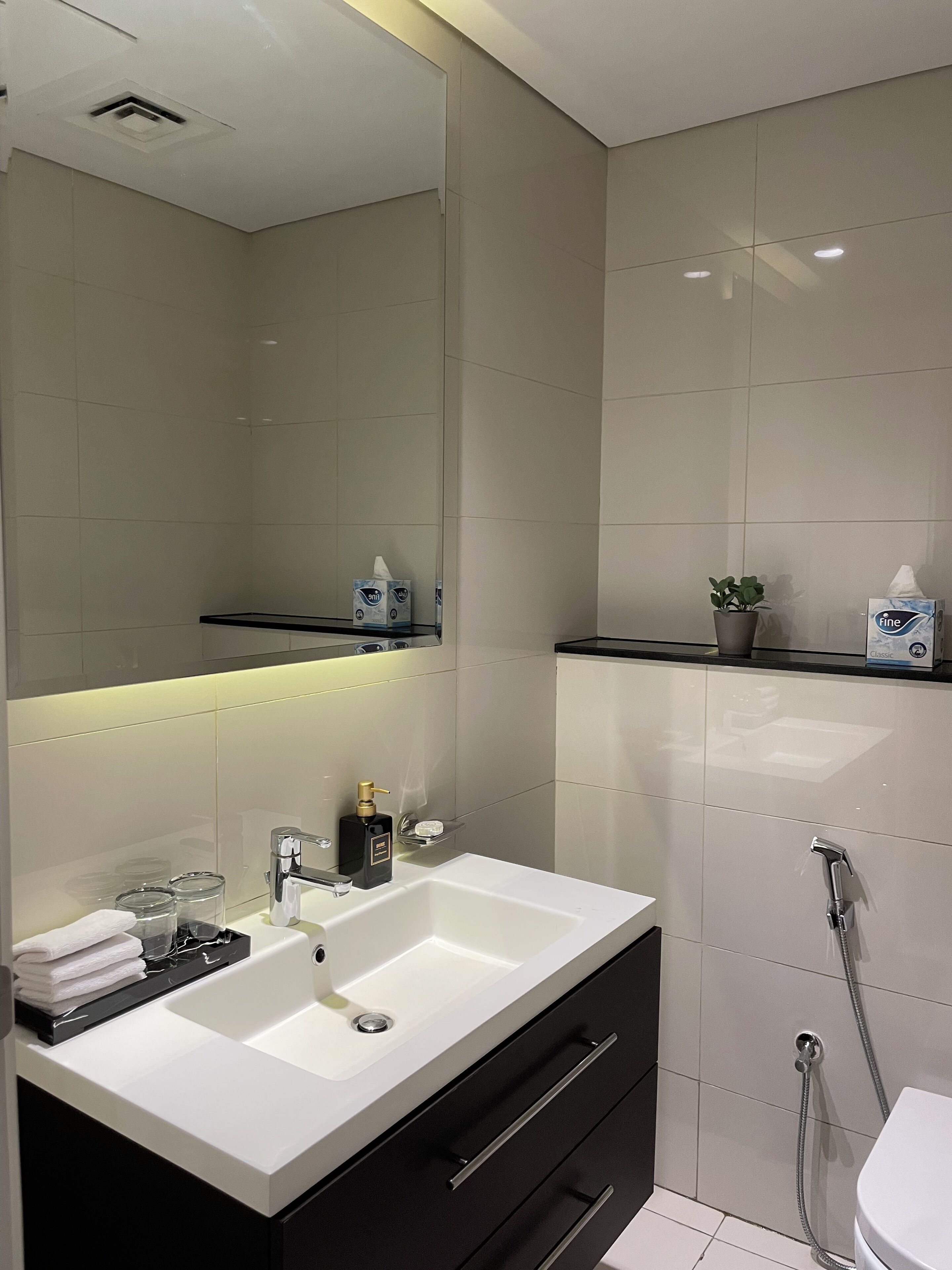 Un baño moderno con un espejo grande, lavamanos blanco, mueble negro y zona de ducha.