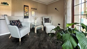 Een moderne woonkamer met twee grijze fauteuils, een glazen salontafel en een groot raam met gordijnen.