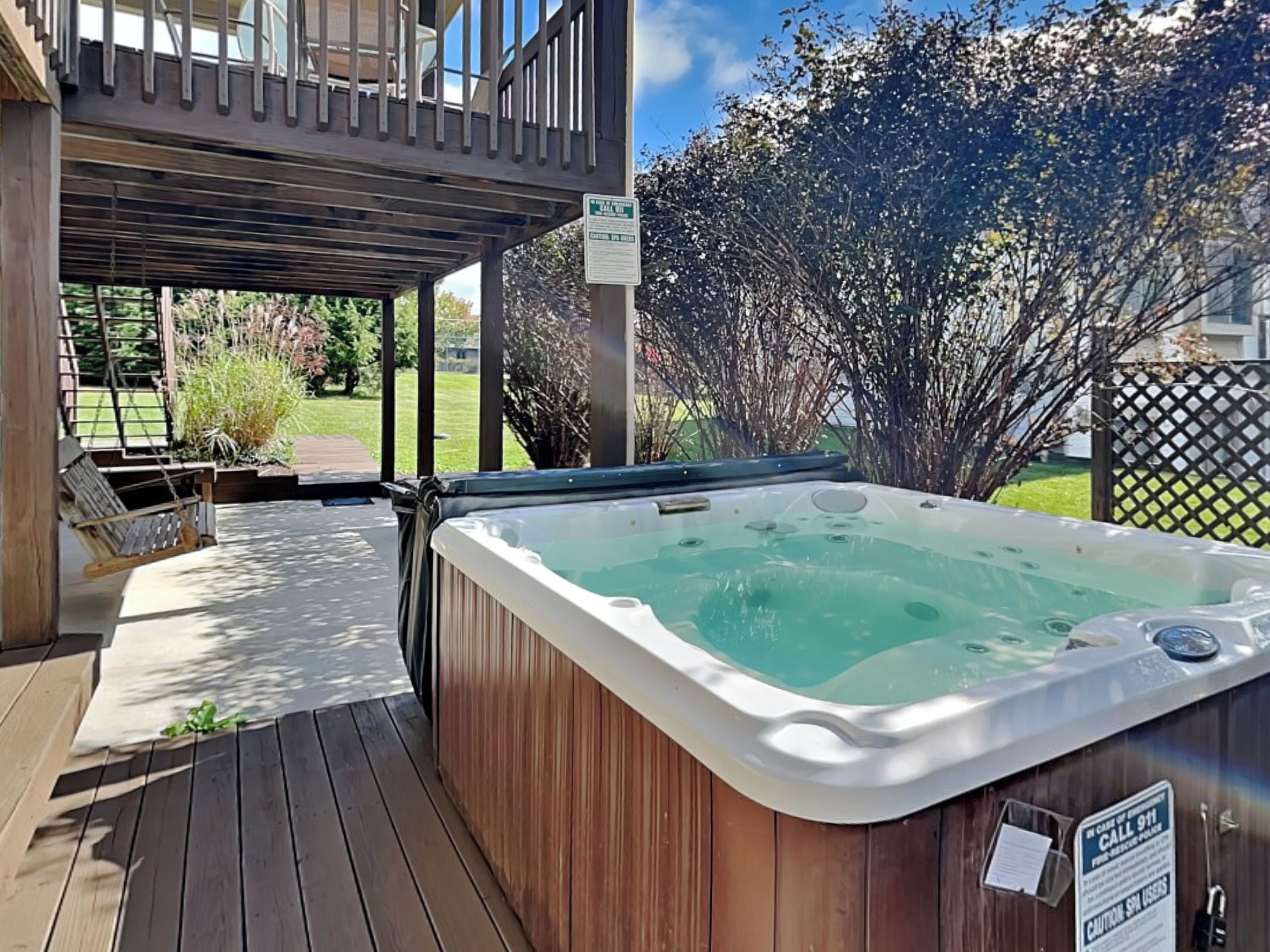 A hot tub on a wooden deck with a covered patio area, surrounded by greenery and a fence.