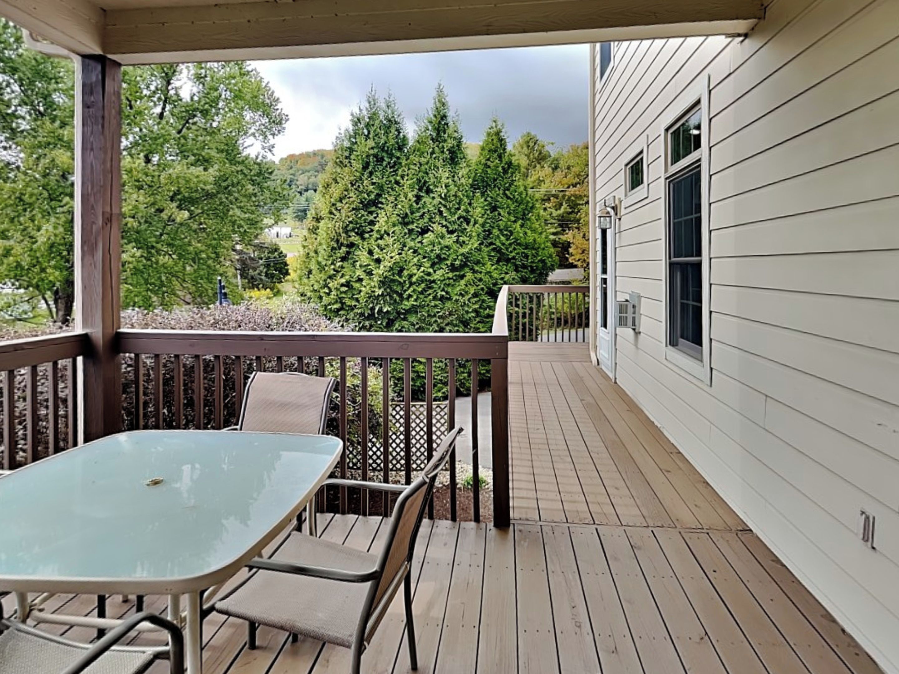 A covered deck with a glass table and chairs, overlooking a wooded area and a house.