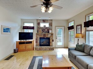 A living room with a stone fireplace, a flat-screen TV, a wooden coffee table, a gray sofa, and a ceiling fan. - Westwind by Taylor Made Deep Creek (McHenry)