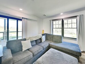 Ein modernes Wohnzimmer mit einer Eckcouch, einem kleinen Tisch und großen Fenstern mit Vorhängen.