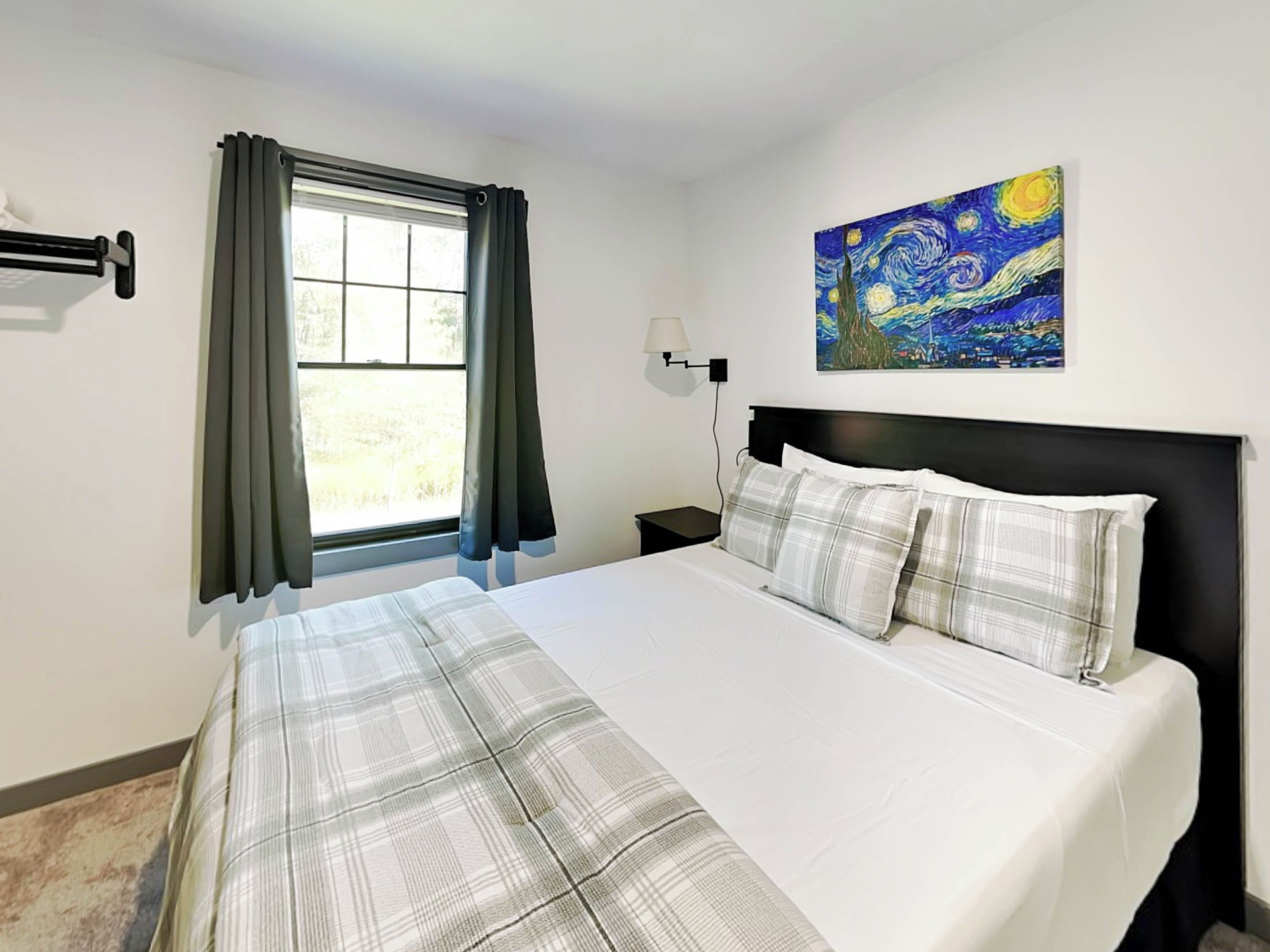 A hotel room with a bed, a window with curtains, a nightstand, a lamp, and a painting on the wall.