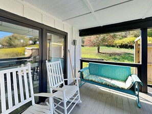 Eine überdachte Veranda mit einem weißen Schaukelstuhl, einer türkisfarbenen Bank mit Blumenkissen und Blick auf einen Garten mit Bäumen und Rasen.