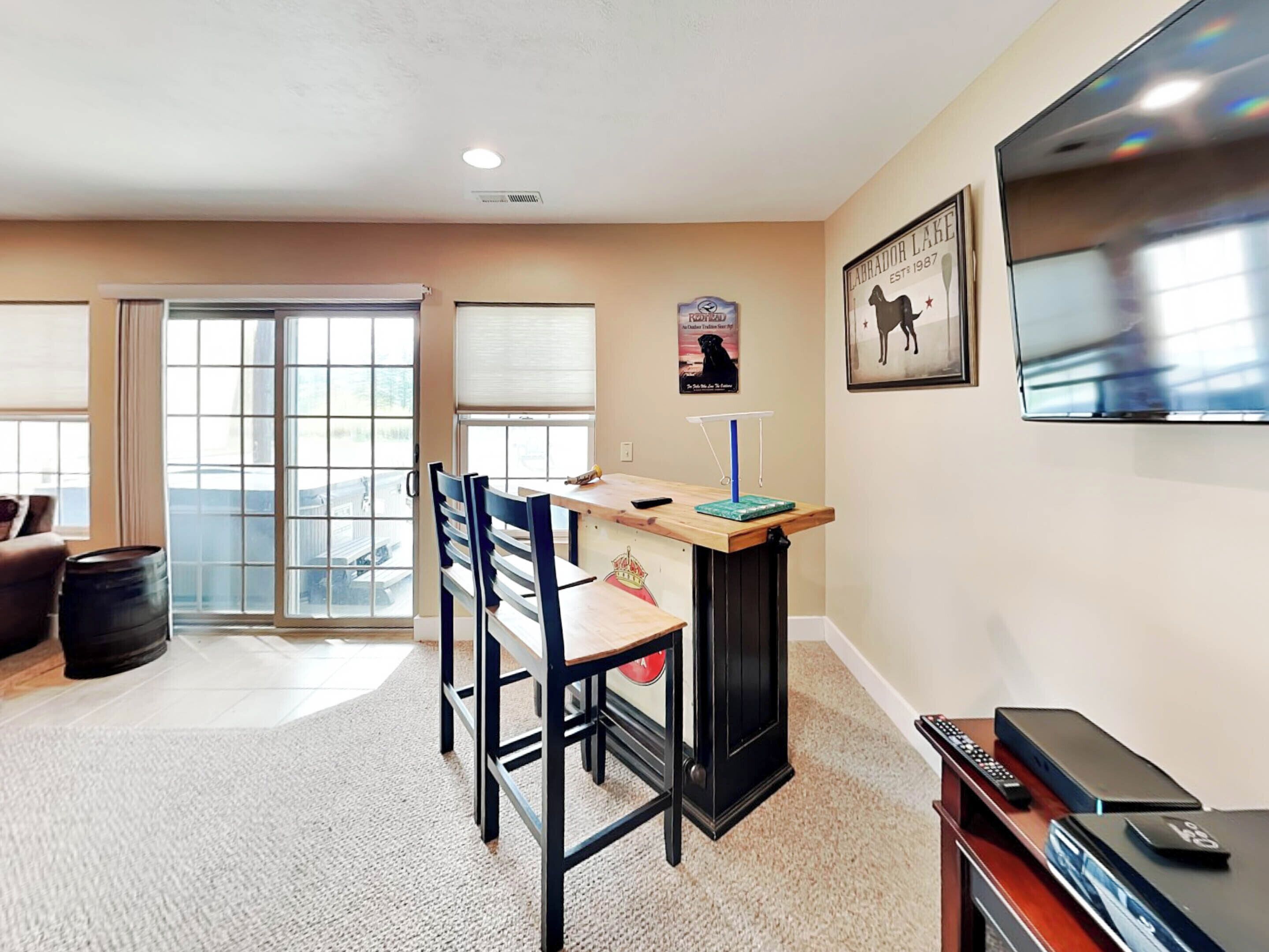 A living room with a dining area, a TV mounted on the wall, and a large window.