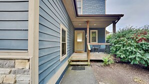 A two-story house with a stone foundation, a front porch with a wooden railing, and a small garden area with shrubs and a tree. - Bluebeary Retreat by Taylor Made Deep Creek (McHenry)