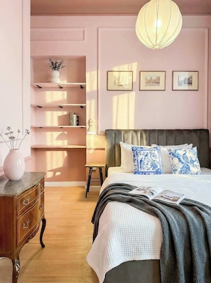 Een slaapkamer met een bed, nachtkastje, lamp en een wandplank met boeken.