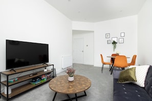 Un salon avec un téléviseur, une table basse ronde, un canapé, une table à manger avec des chaises, et une porte blanche en arrière-plan.