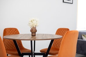 A round wooden table with a vase of white flowers, surrounded by four orange chairs with black frames.