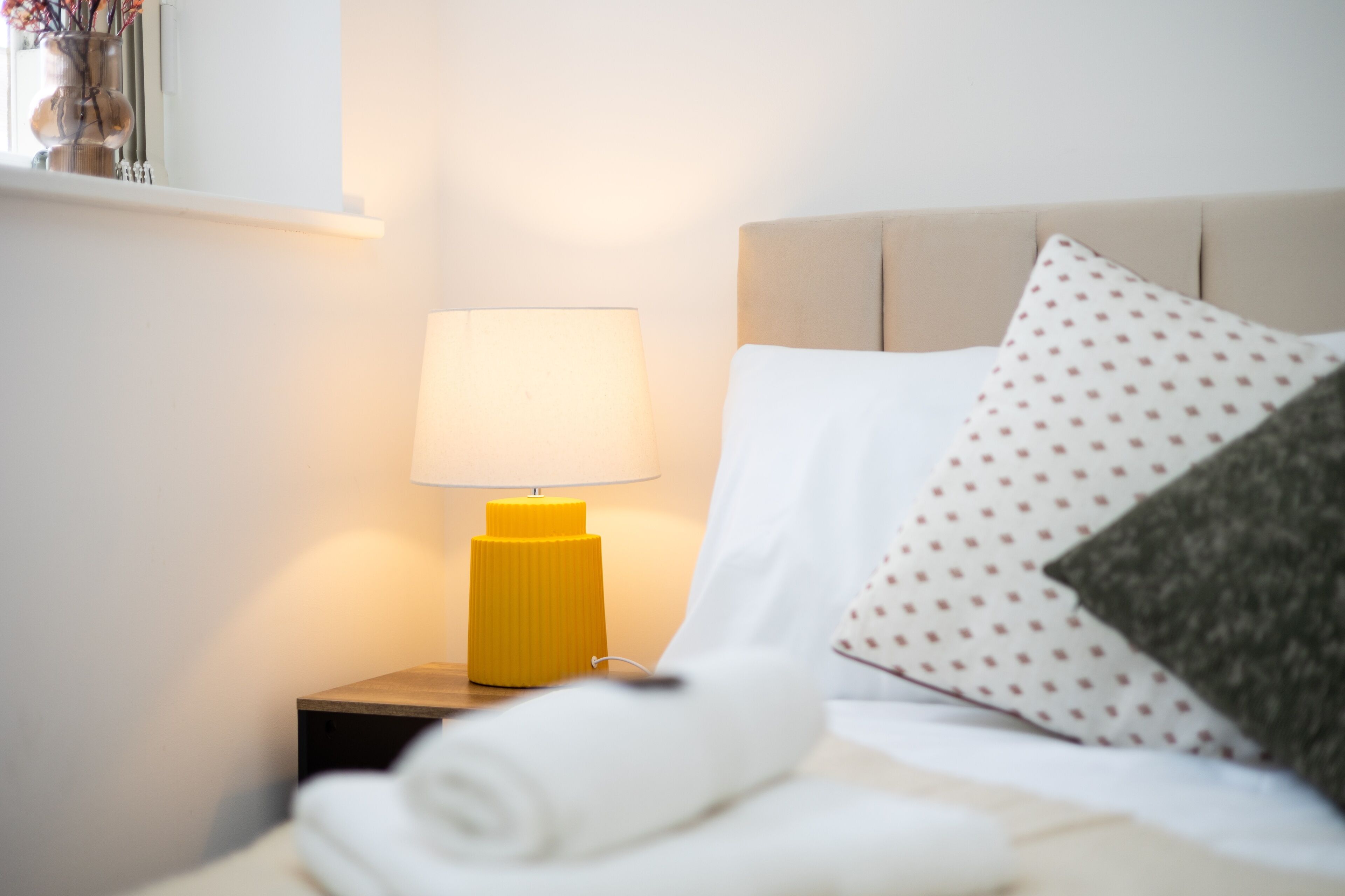 Una cama bien tendida con sábanas blancas, una toalla enrollada, una lámpara de noche y un cojín estampado con lunares.