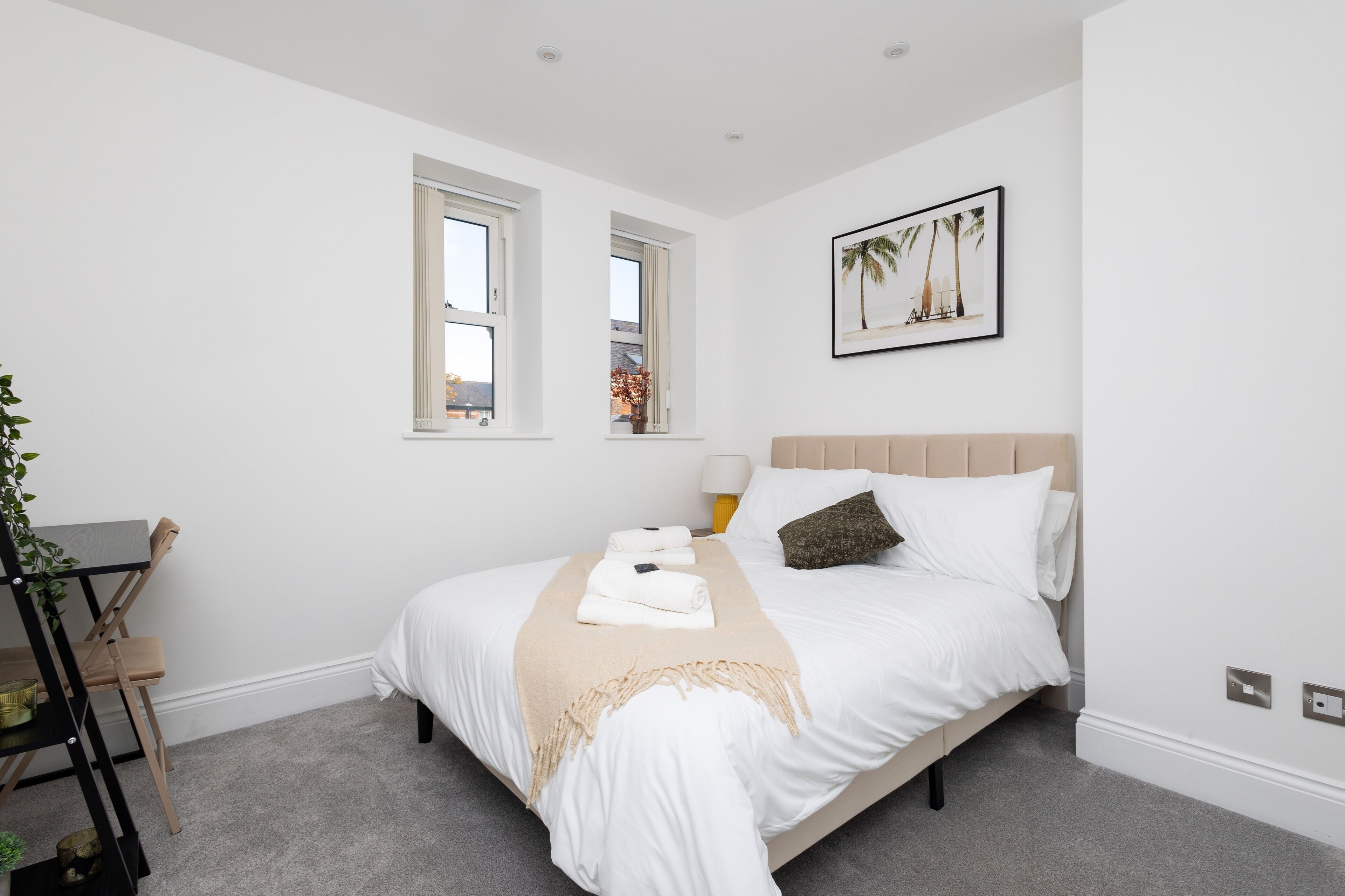 A neatly arranged bedroom with a bed, two windows, a bedside table with a lamp, and a framed picture on the wall.