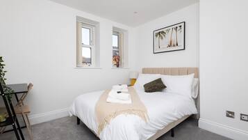 A neatly arranged bedroom with a bed, two windows, a bedside table with a lamp, and a framed picture on the wall.