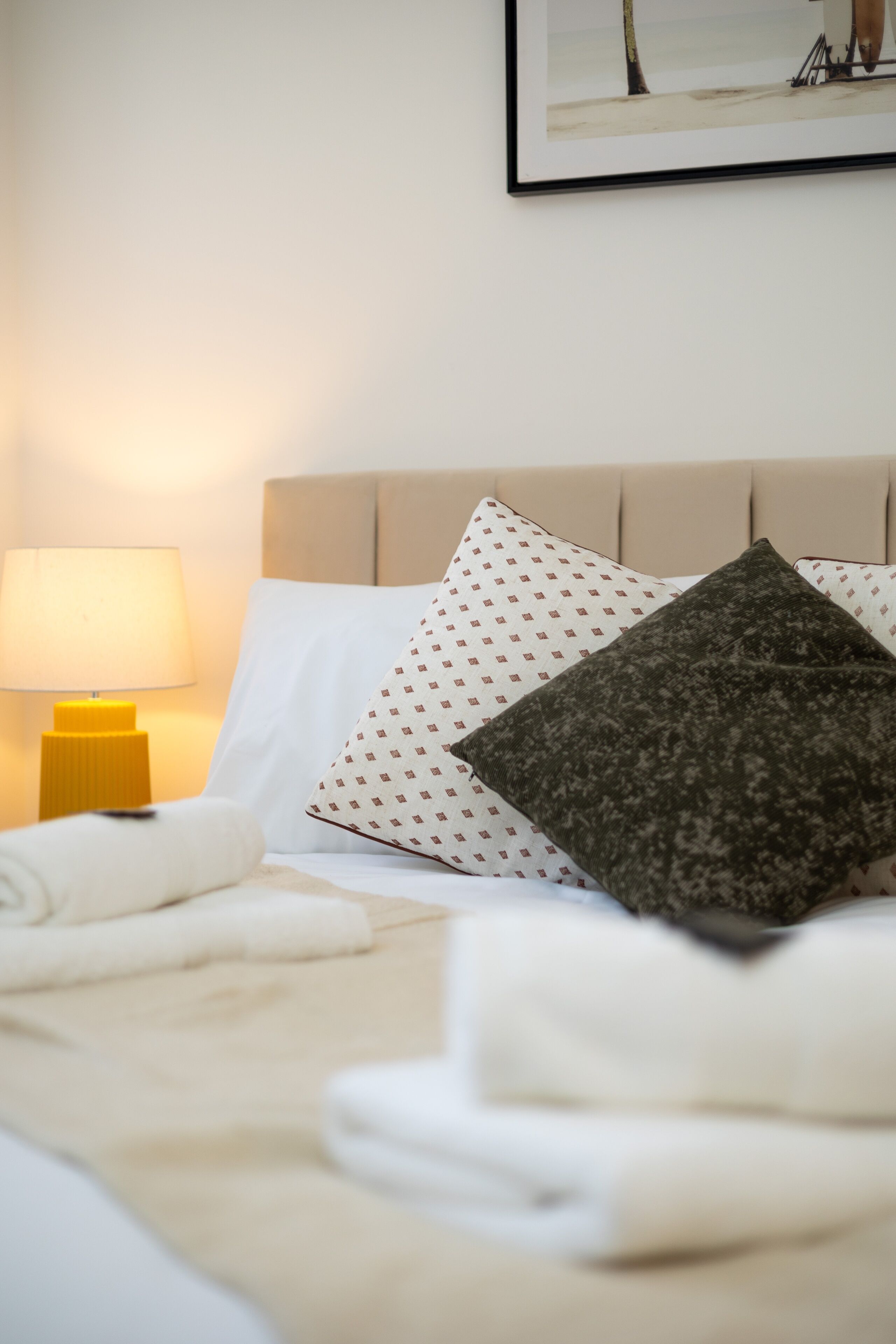 Un lit bien fait, avec des oreillers blancs et à motifs, une lampe de chevet jaune et un tableau encadré accroché au mur.