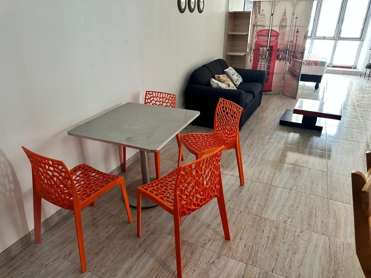 A dining area with a table and four orange chairs, a black sofa, and a red telephone booth decoration on the wall.