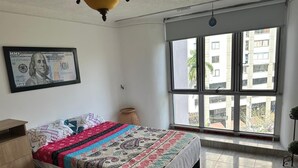Un dormitorio con una cama grande, una ventana con vistas a edificios, un ventilador de techo y un billete de dólar enmarcado en la pared.