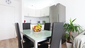 Une cuisine de taille réduite avec des meubles blancs, un four à micro-ondes et un réfrigérateur. Un panier de fruits est posé sur la table, et une plante en pot se trouve près de la fenêtre.