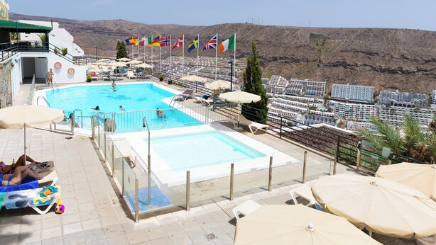 Une piscine dotée de chaises longues et de parasols, de drapeaux, et offrant une vue sur les bâtiments et les collines.
