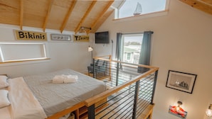 Une chambre avec un lit superposé, un plafond en bois, une fenêtre donnant sur un beau paysage, et une enseigne indiquant « Maillots de bain » et « T-shirts ».