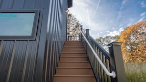 Un escalier en bois menant à un bâtiment doté d’une rambarde en métal et d’une fenêtre sur laquelle on peut lire une inscription.